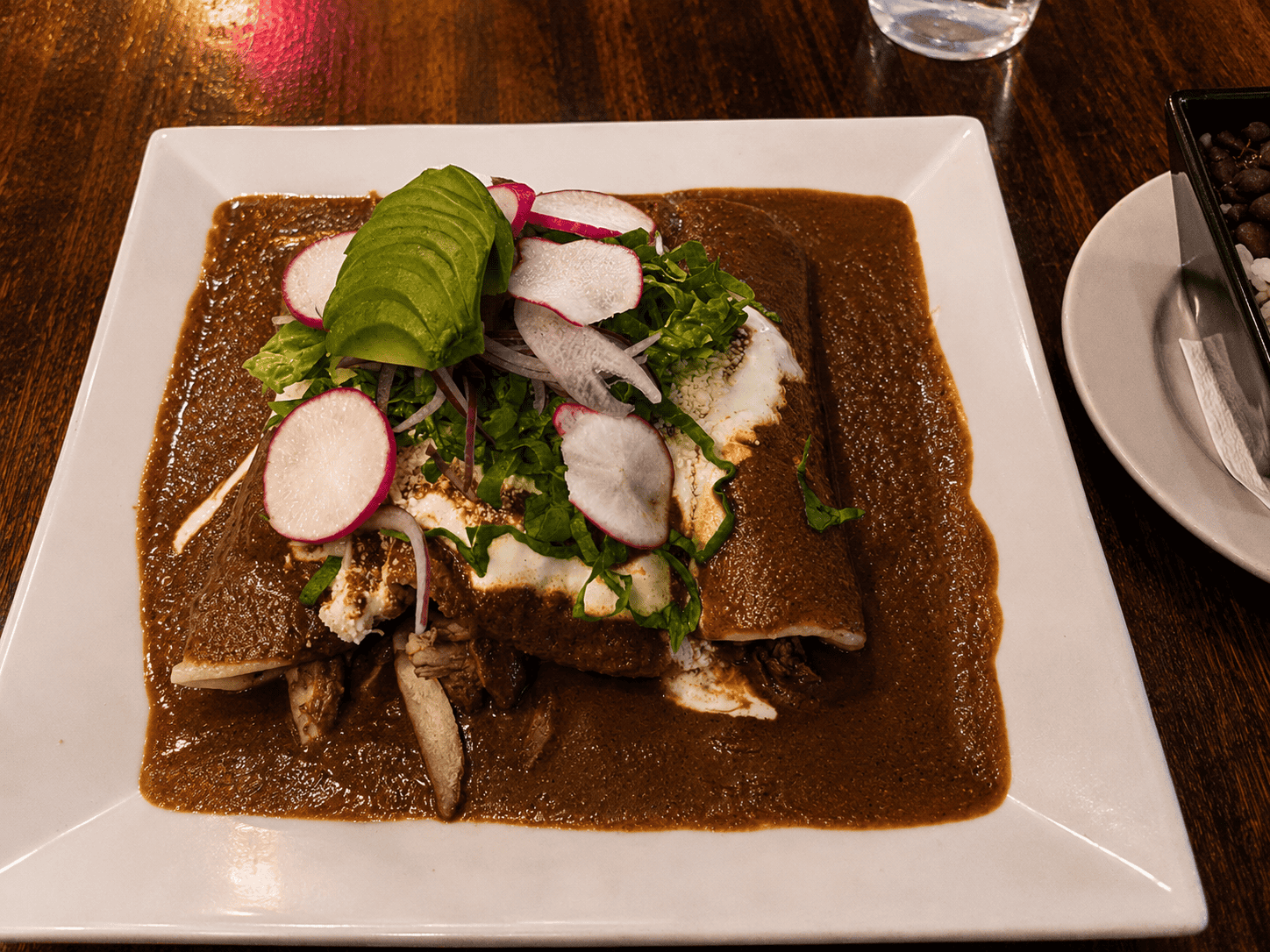 Authentic Mexican chicken enchiladas covered in rich dark mole sauce topped with sliced avocado, radishes, and crema