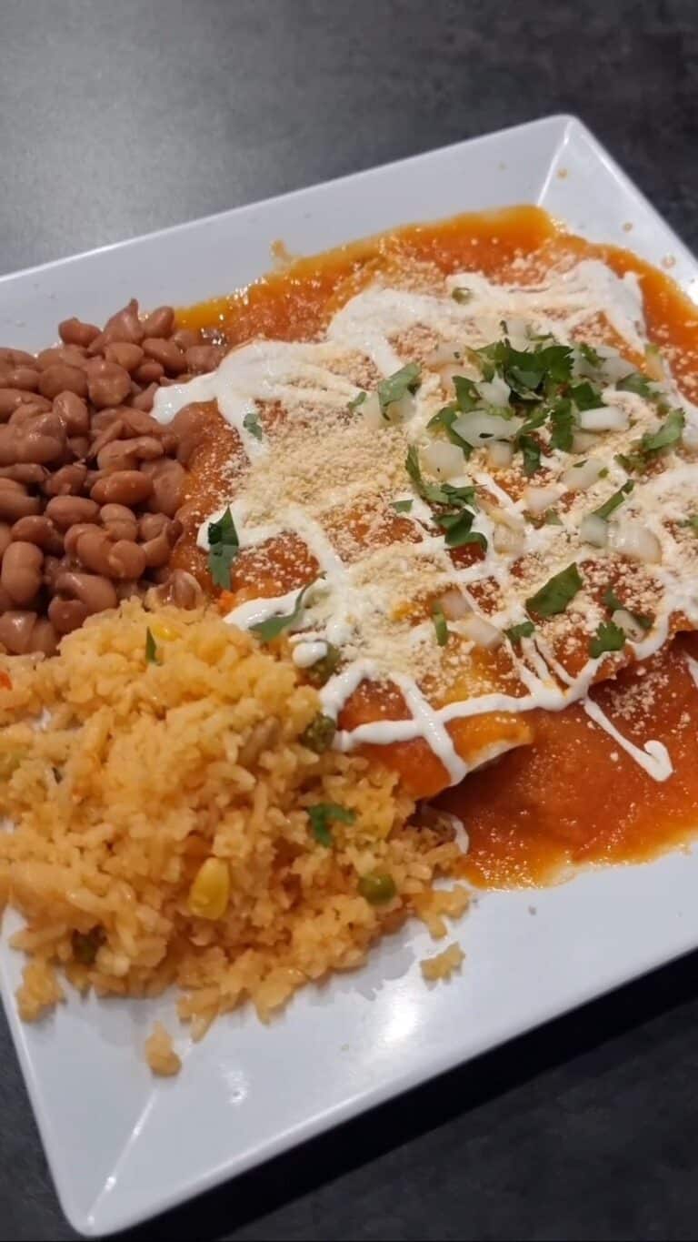 A plate of chicken enchiladas smothered in red tomato sauce, melted cheese, and sour cream drizzle, served with sides of Mexican rice and pinto beans.