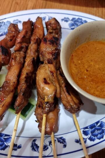 Grilled Malaysian chicken satay skewers served on a blue floral plate with a side bowl of thick peanut dipping sauce.
