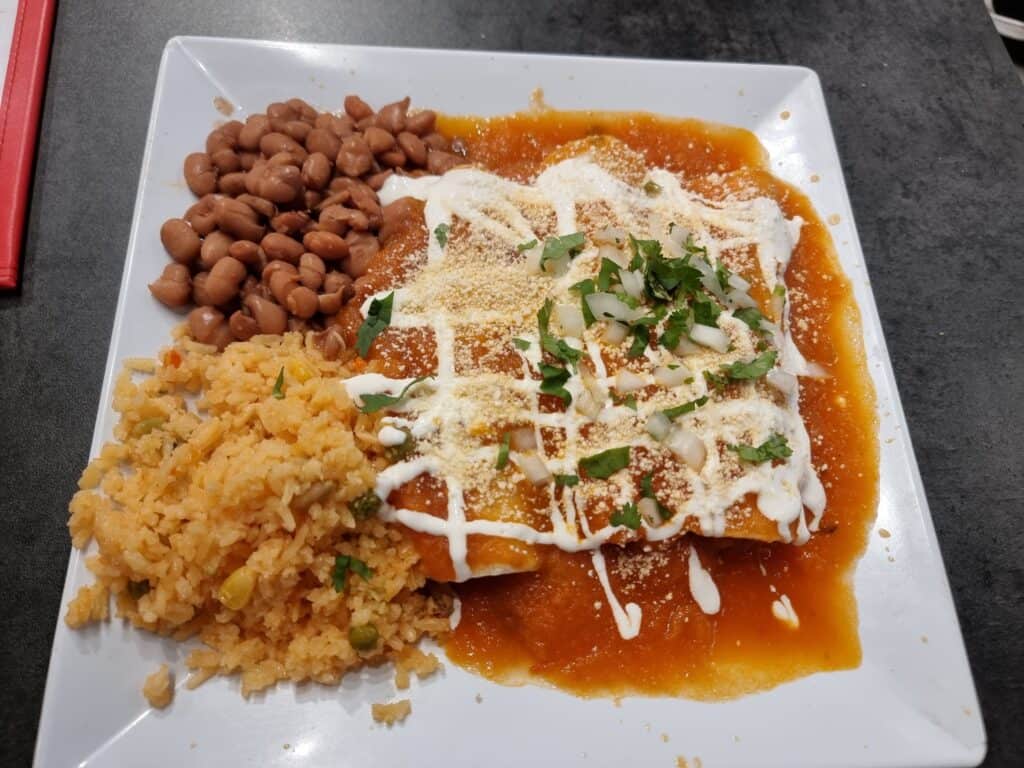 A plate of chicken enchiladas smothered in red tomato sauce, melted cheese, and sour cream drizzle, served with sides of Mexican rice and pinto beans.