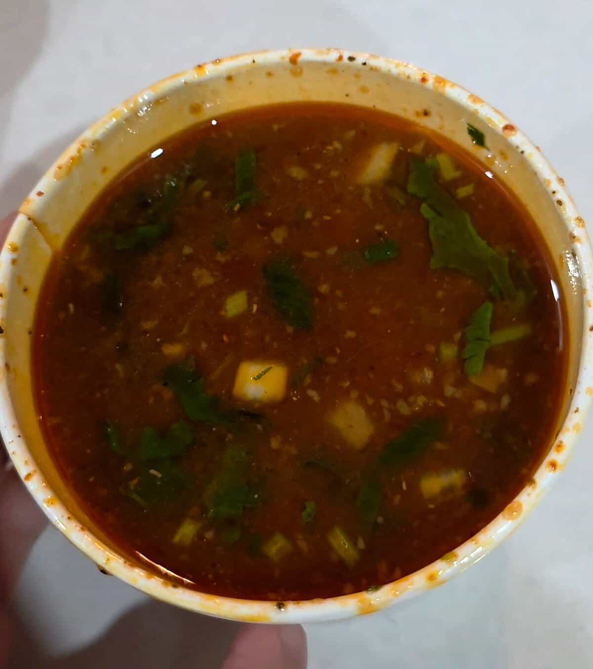 A overhead view of a paper cup filled with rich, dark red birria consommé broth topped with fresh cilantro and chopped onions.