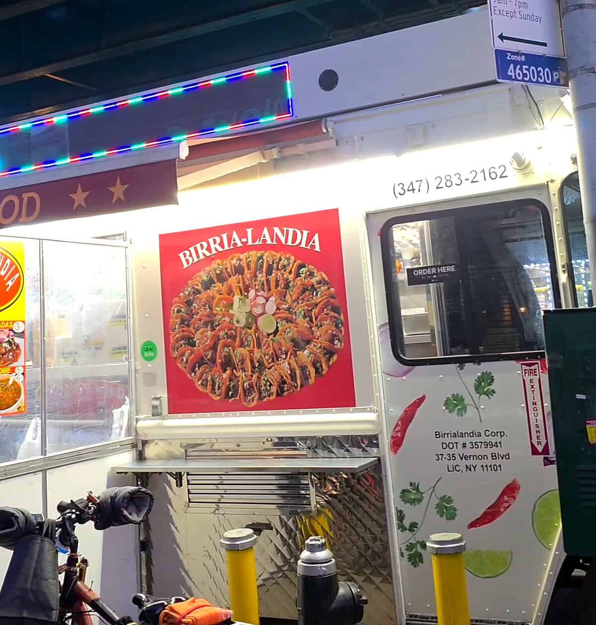 The exterior of the famous Birria-Landia food truck parked at night under the subway tracks in Jackson Heights, Queens.