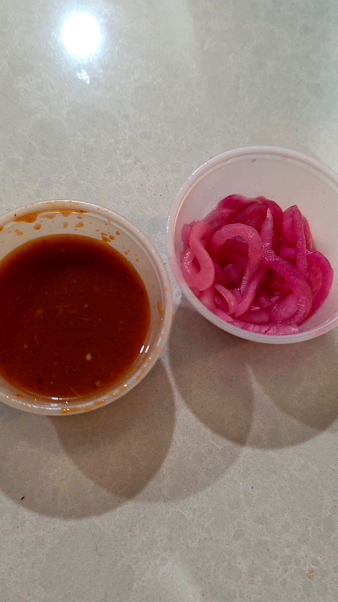Two small plastic condiment cups containing bright pink pickled onions and a dark red spicy salsa on a counter.