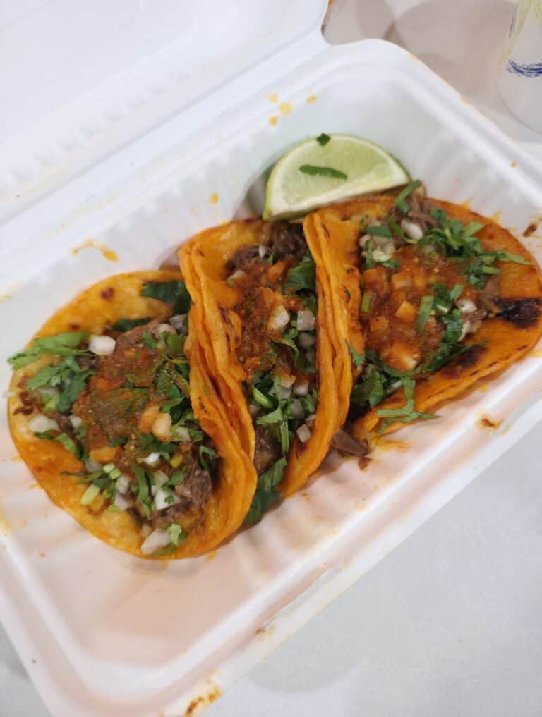 Three crispy Birria-Landia tacos topped with fresh chopped onions, cilantro, and red salsa, served with a lime wedge in a takeout container.