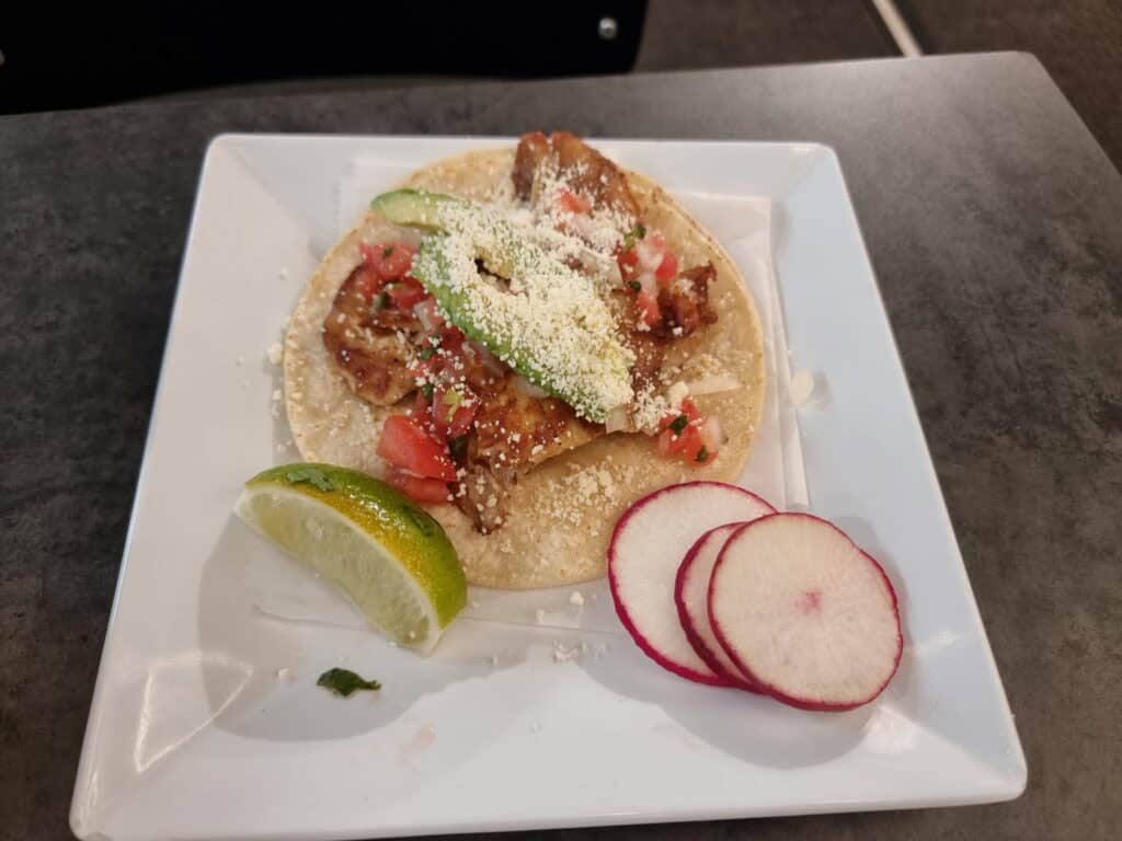 A single authentic chicken taco on a corn tortilla topped with avocado slices, cotija cheese, pico de gallo, served with radish slices and a lime wedge.