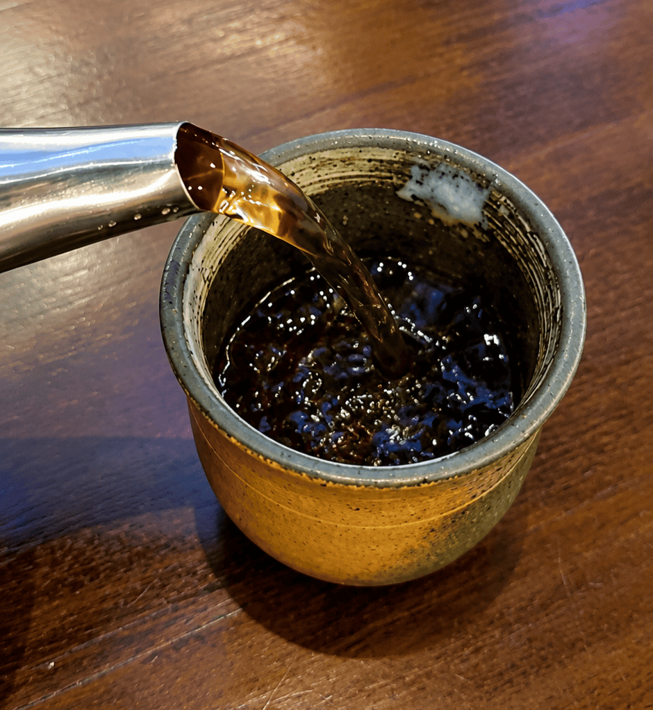 Warm hot tea being poured from a metal kettle into a traditional ceramic tea cup on a dark wood table.