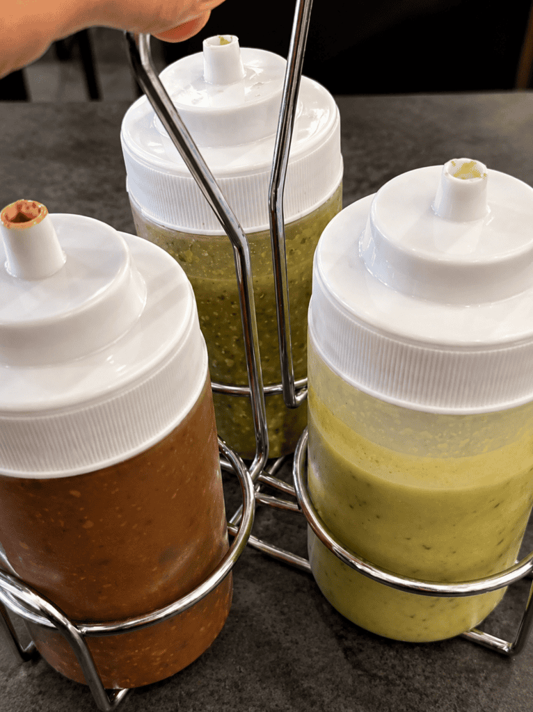 Three squeeze bottles filled with guacamole sauce, salsa verde green sauce, and red hot sauce in a wire caddy.