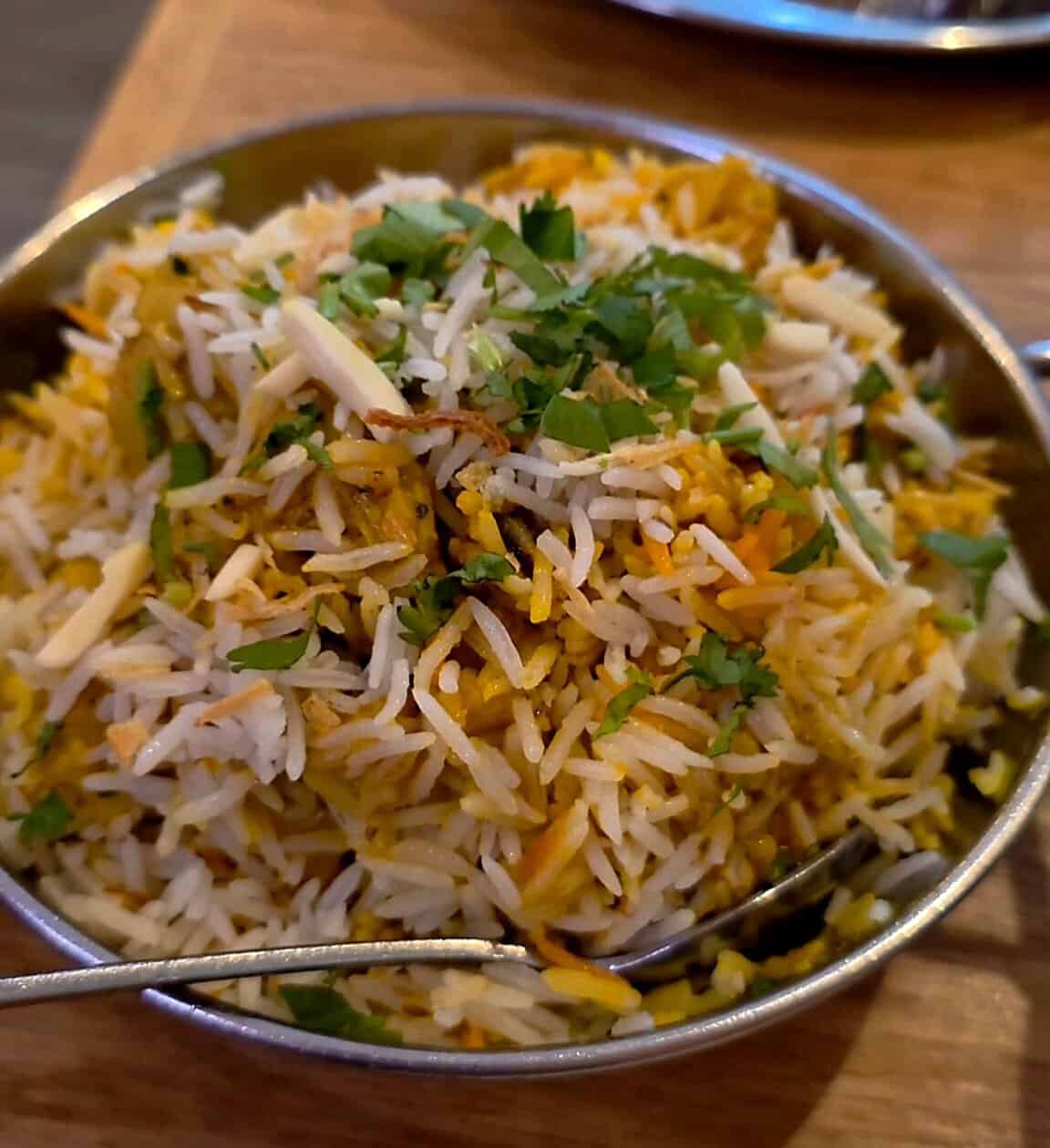 A metal bowl filled with aromatic chicken biryani rice mixed with spices and garnished with slivered almonds and fresh cilantro.