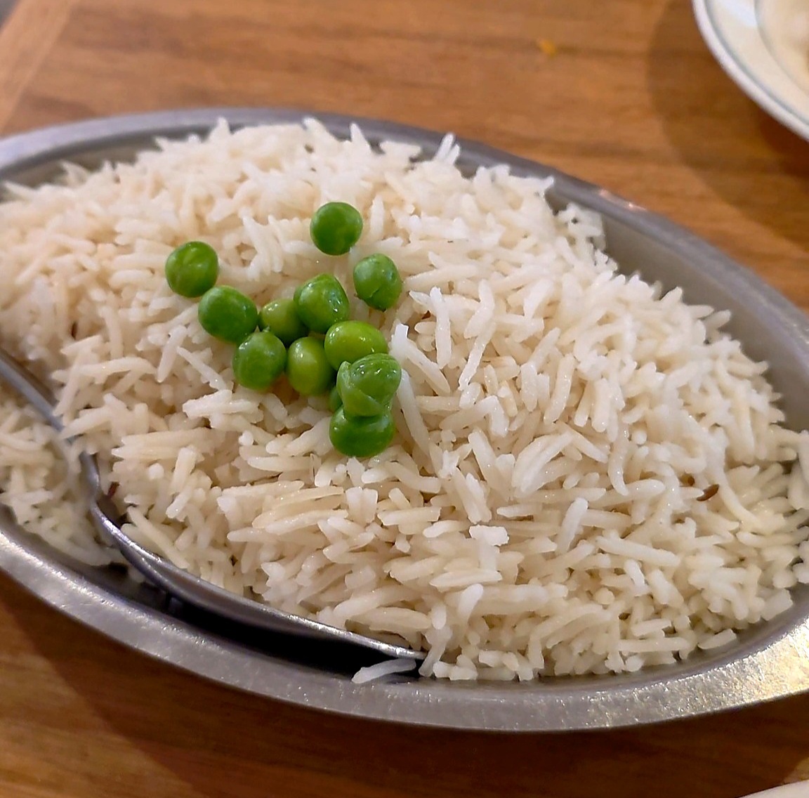 An oval metal serving dish filled with steaming hot, long-grain white basmati rice decorated with a small pile of green peas on top.