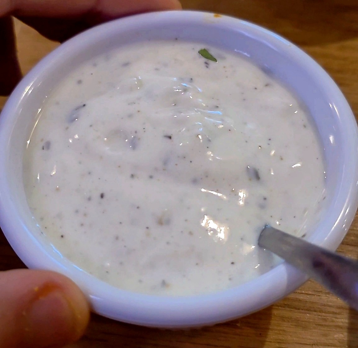 A small white ceramic bowl containing smooth, creamy Indian garlic yogurt sauce mixed with fine black pepper and spices.