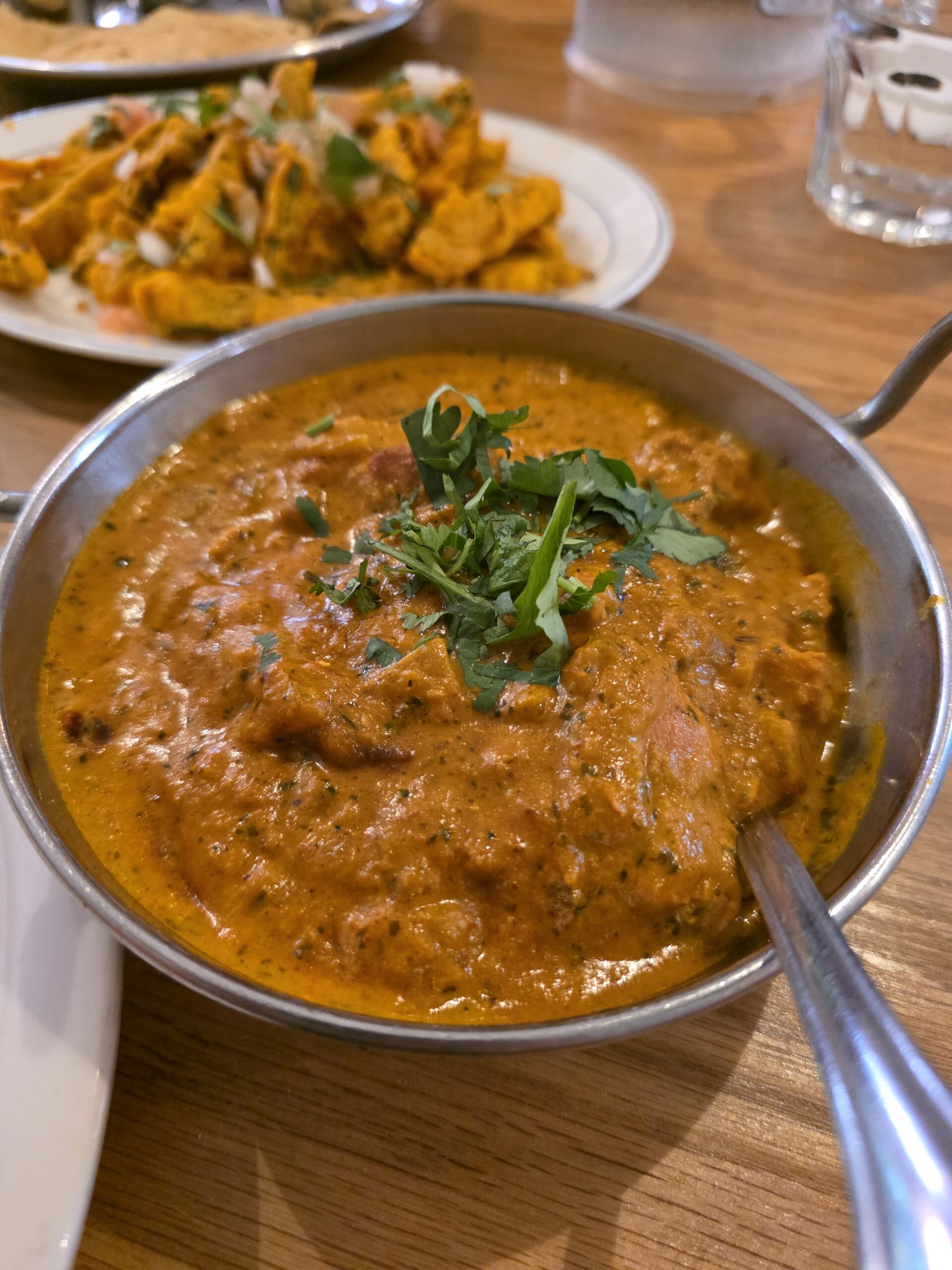 A close-up view of rich, vibrant orange chicken tikka masala curry in a stainless steel balti bowl garnished with chopped cilantro.