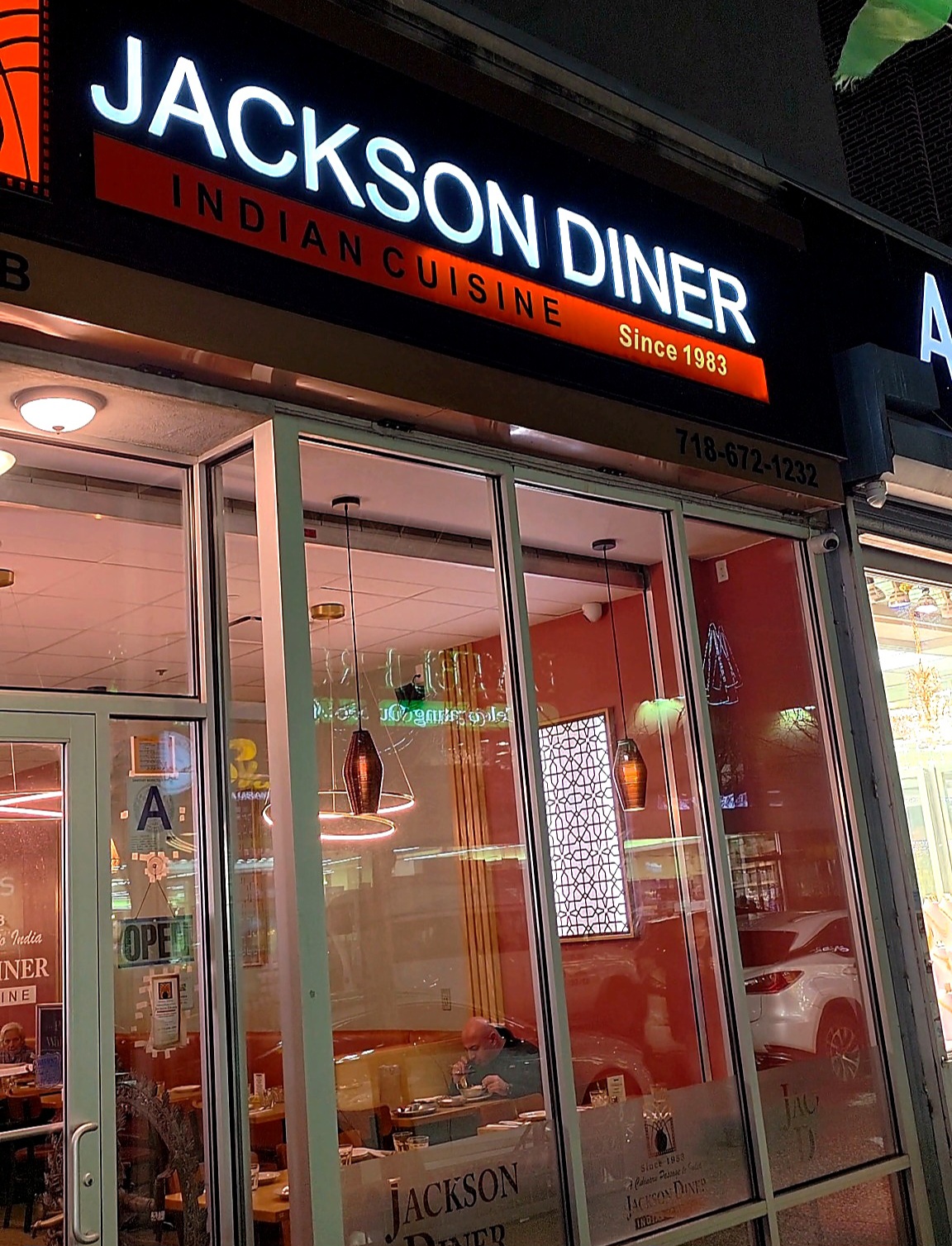 The vibrant neon sign and glass storefront window of Jackson Diner Indian Cuisine at night on 74th Street in Jackson Heights, Queens.