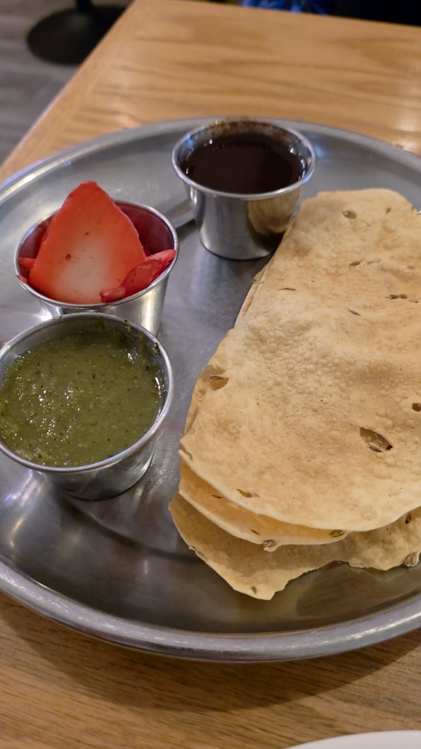 A silver platter serving crispy Indian papadum crackers alongside three small metal cups containing green chutney, dark tamarind sauce, and red pickle.