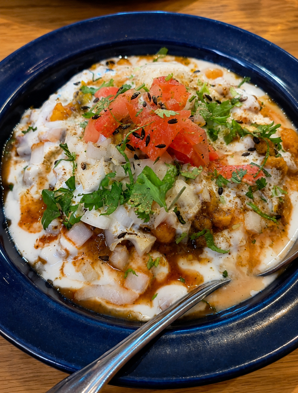 A close-up view of a colorful bowl of Samosa Chaat at Jackson Diner, layered with creamy white yogurt, chickpea curry, diced raw onions, chopped tomatoes, and fresh cilantro.