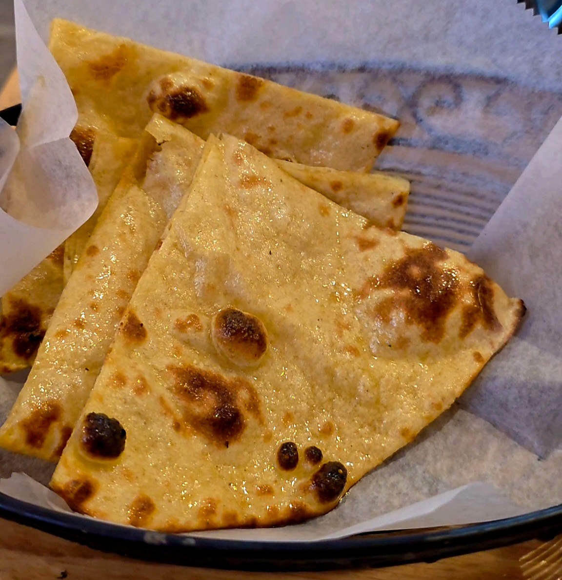 Several triangular slices of fresh, glistening butter naan bread with charred bubbles sitting inside a basket lined with white paper.