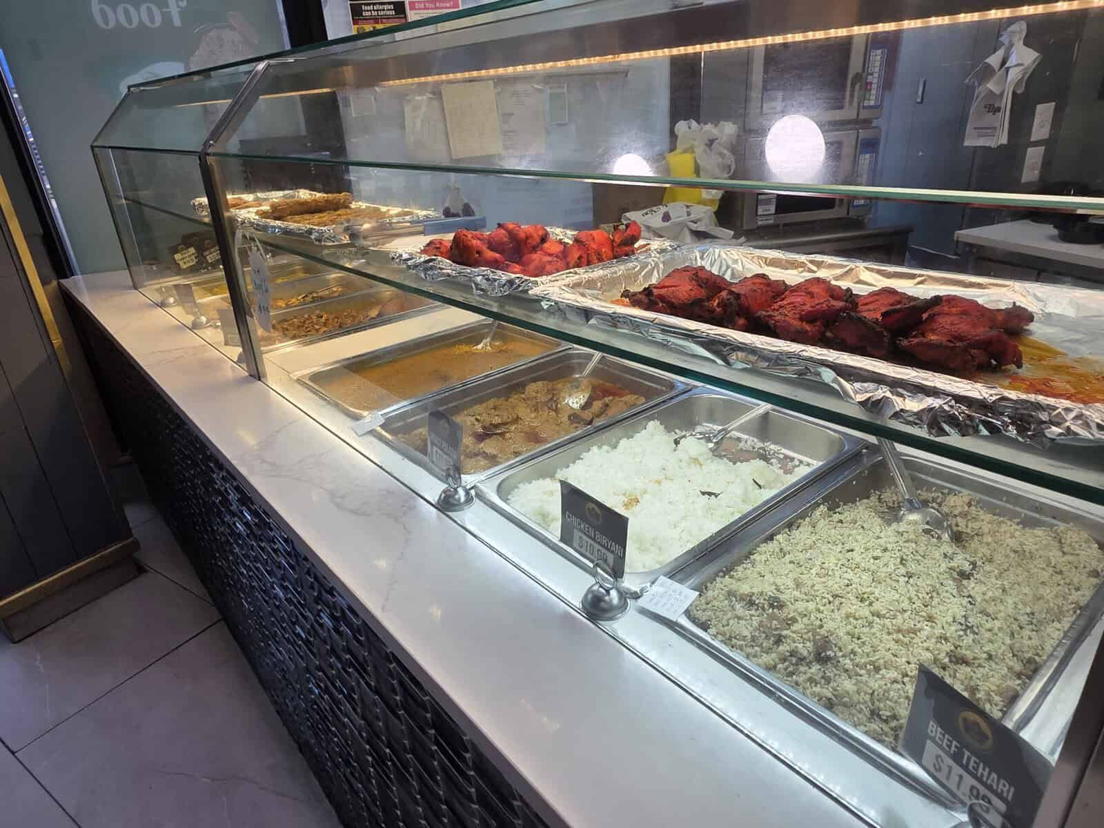 A glass display counter at Khalil Biryani House showing pre-made Bangladeshi dishes including Tandoori chicken, beef tehari, rice, and curries in metal trays.
