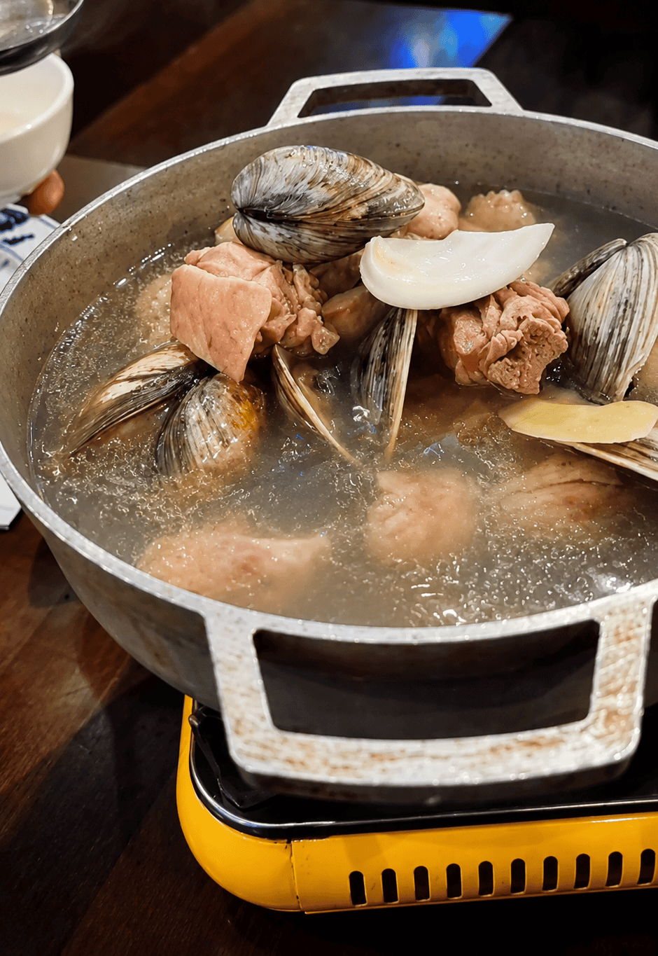 Clam and chicken soup with fresh ginger simmering in a metal pot on a tabletop burner.