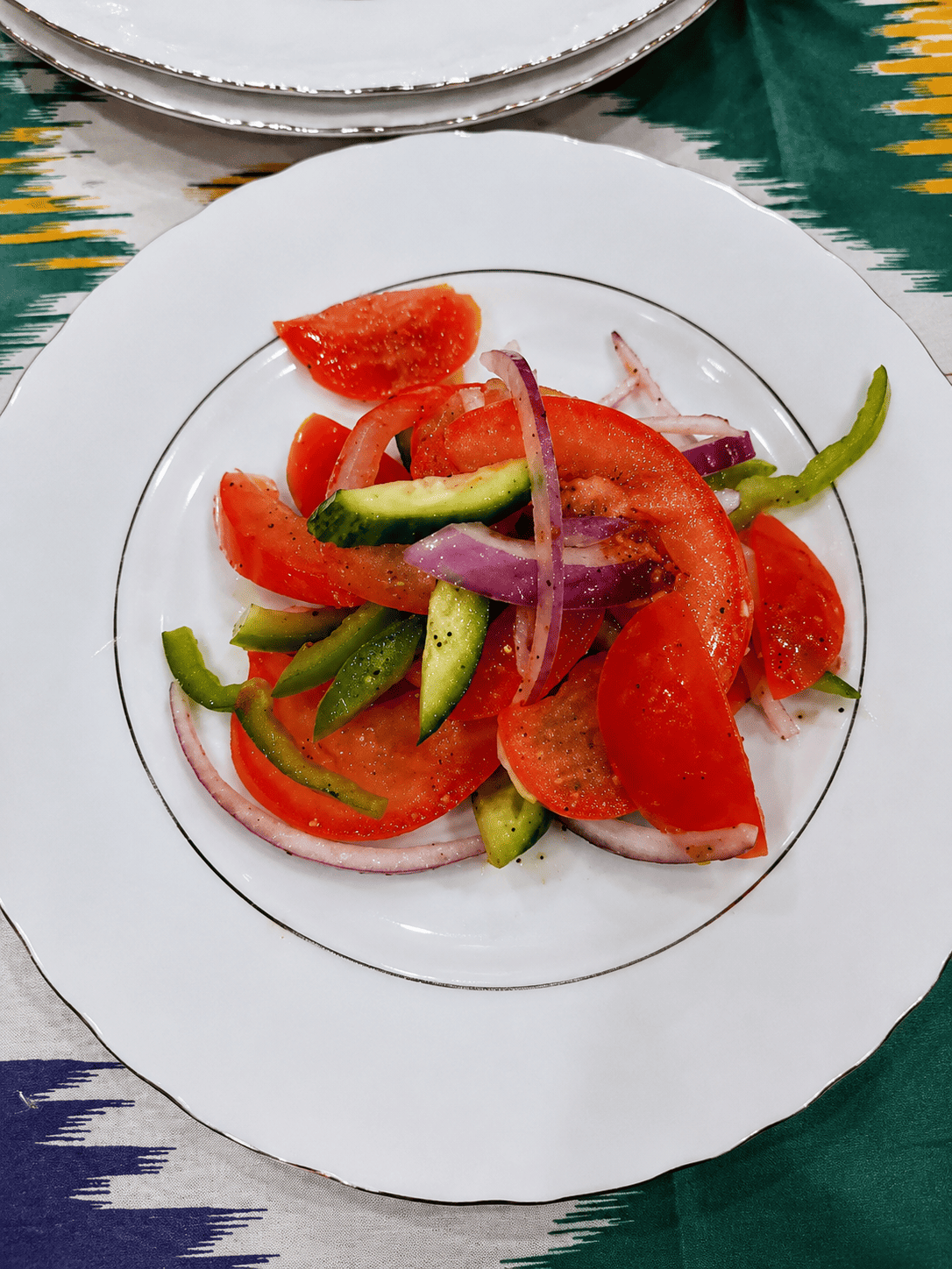  A clean white plate presenting a fresh Uzbek salad made with sliced raw red onions, cucumbers, green peppers, and juicy red tomatoes seasoned with coarse black pepper.