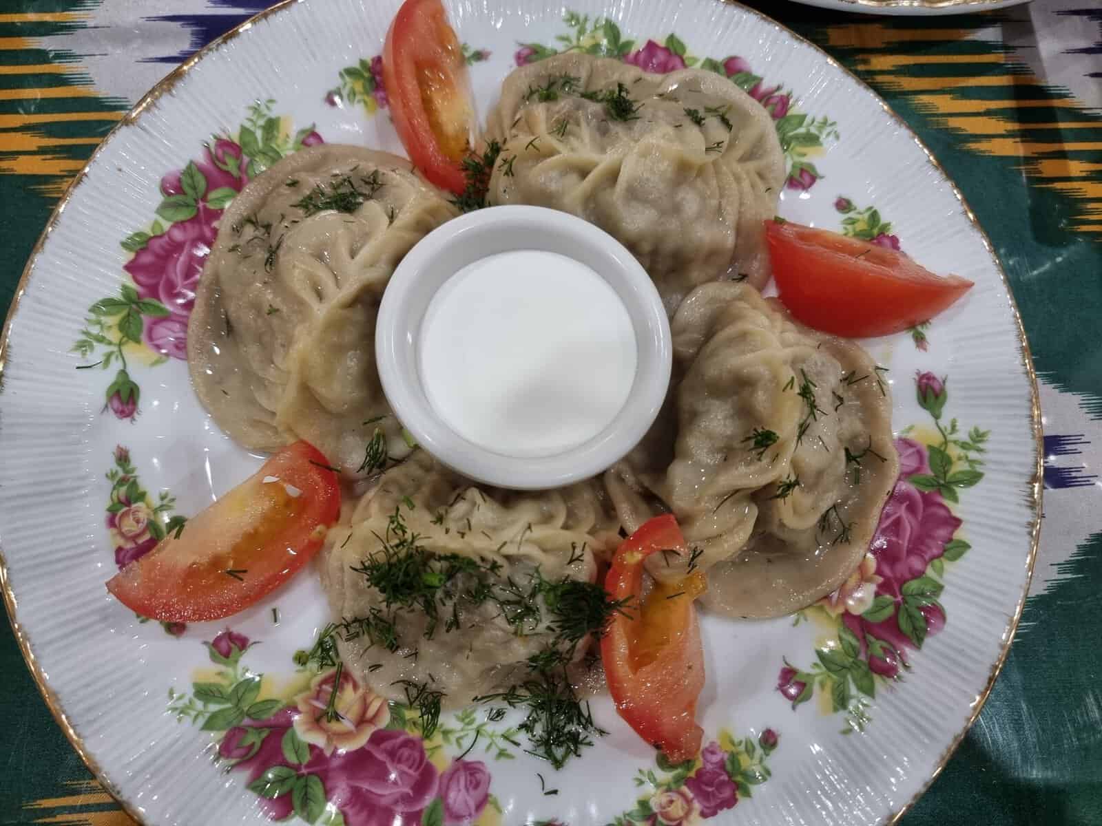  A floral pattern plate holding four large steamed beef Manti dumplings garnished with fresh dill, served with fresh tomato wedges and a center cup of tangy white yogurt sauce.
