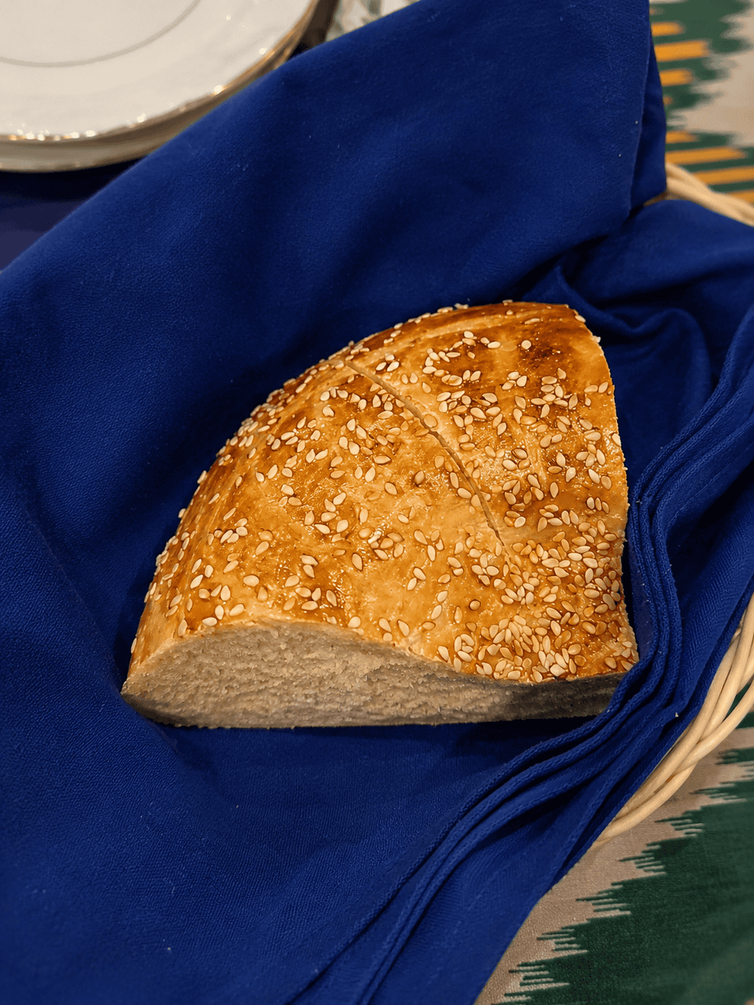 A thick wedge of golden-brown traditional Uzbek bread topped with sesame seeds sitting inside a basket lined with a vibrant blue cloth napkin.