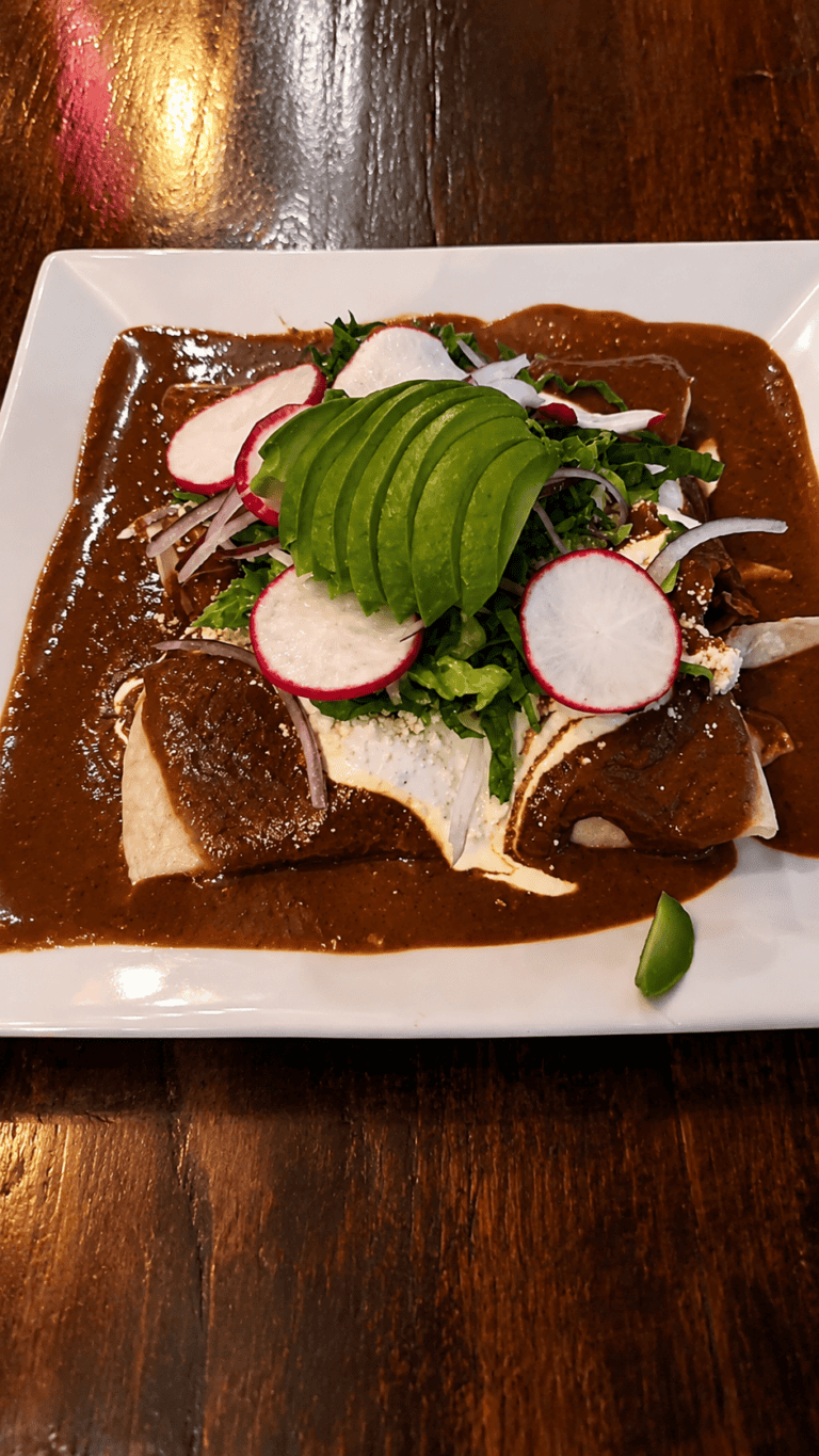Authentic Mexican chicken enchiladas covered in rich dark mole sauce topped with sliced avocado, radishes, and crema