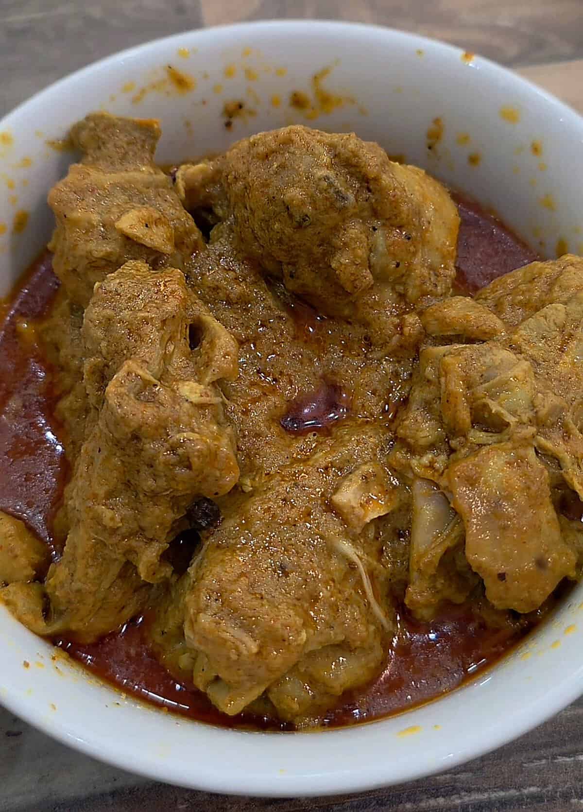A white ceramic bowl filled with rich, oil-glistening South Asian chicken curry in a deeply spiced red gravy.