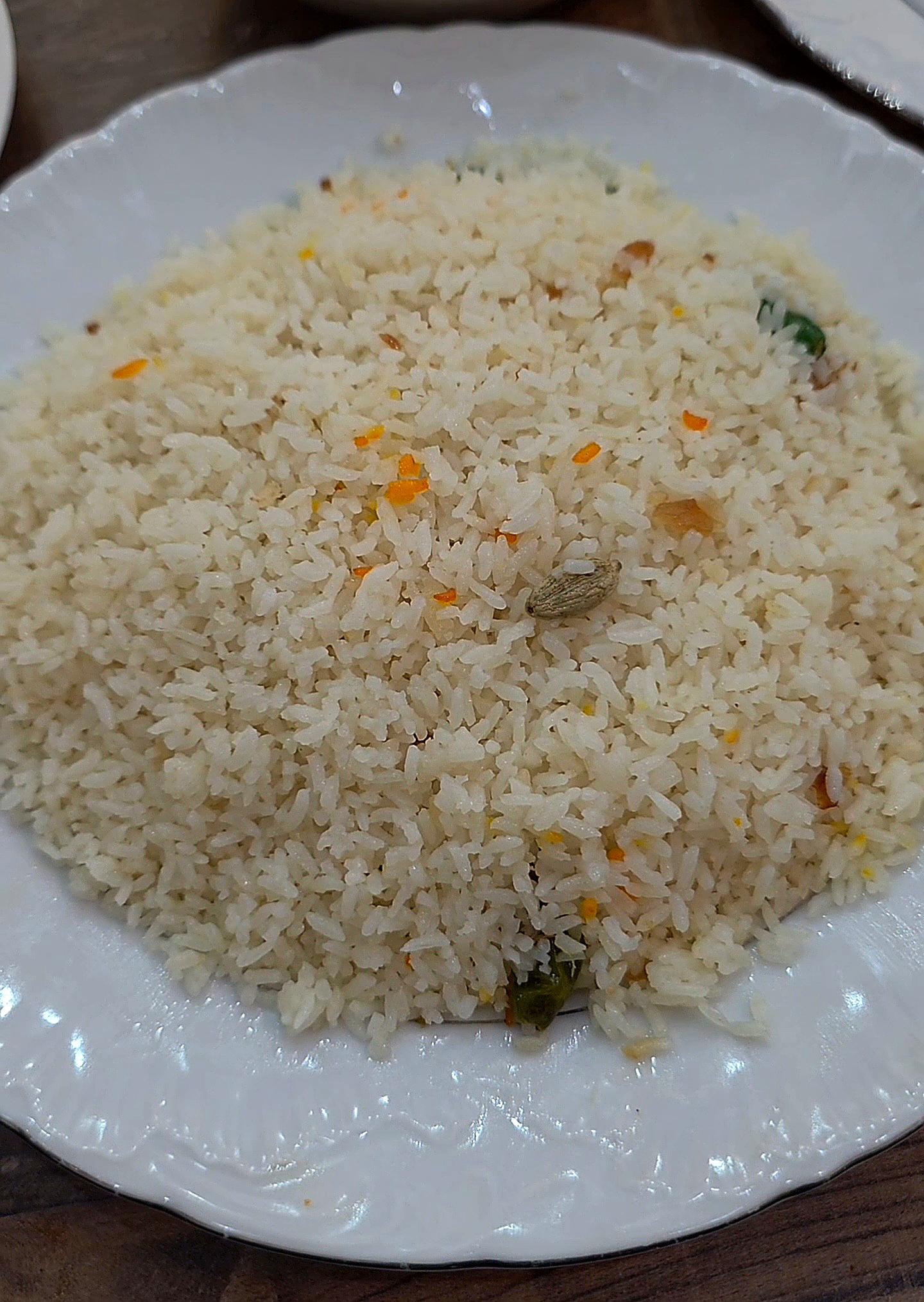 A mound of fluffy, aromatic white pulao rice garnished with orange food coloring highlights and whole spices on a white plate.