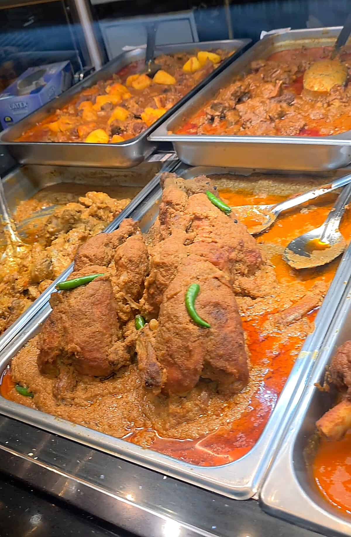 Metal steam trays loaded with traditional South Asian curries and roasted whole chicken dishes behind a glass counter at Shanai Restaurant.