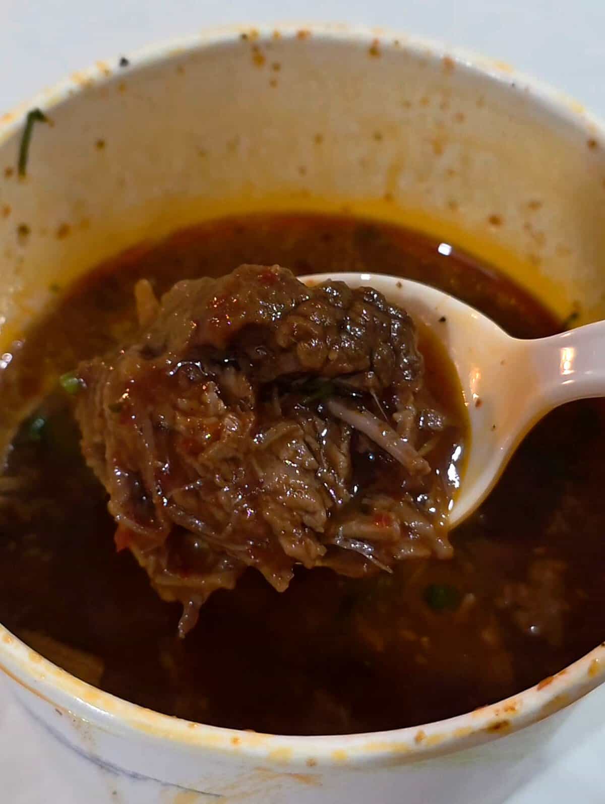 A white plastic spoon lifting a tender, juicy piece of slow-cooked shredded birria beef out of a cup of flavorful consommé broth.