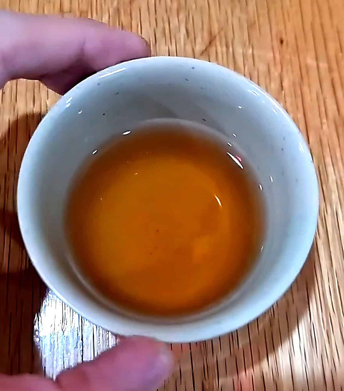 Overhead view of a hand holding a small ceramic cup filled with warm, golden complimentary hot tea on a wooden table.