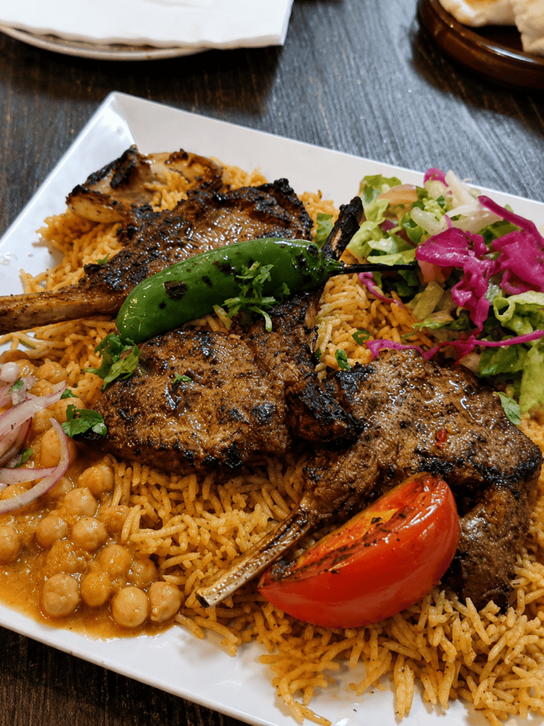 A heavily loaded platter of grilled lamb chops over orange seasoned rice, served with chickpeas, a charred green pepper, a grilled tomato, and a red cabbage side salad.