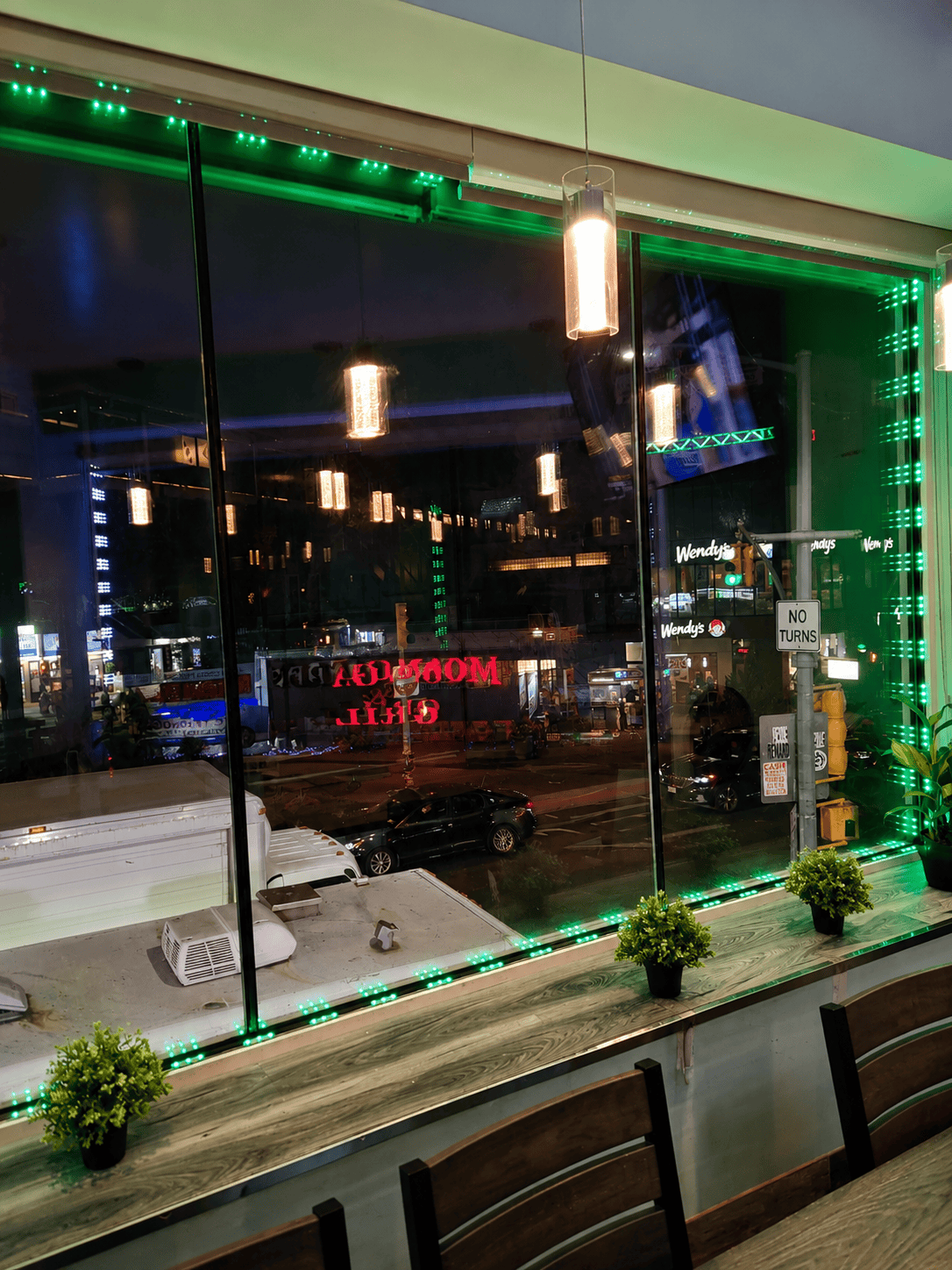 The casual second-floor dining area of Moonlite Grill and Chicken at night, featuring large glass windows overlooking Broadway with green LED accent lighting.