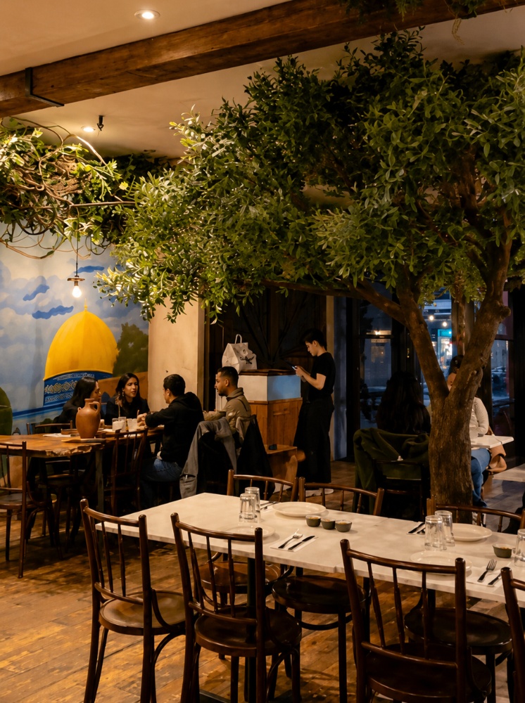 The cozy interior of Al Badawi restaurant in Brooklyn with a large olive tree and a mural of the Dome of the Rock on the wall.