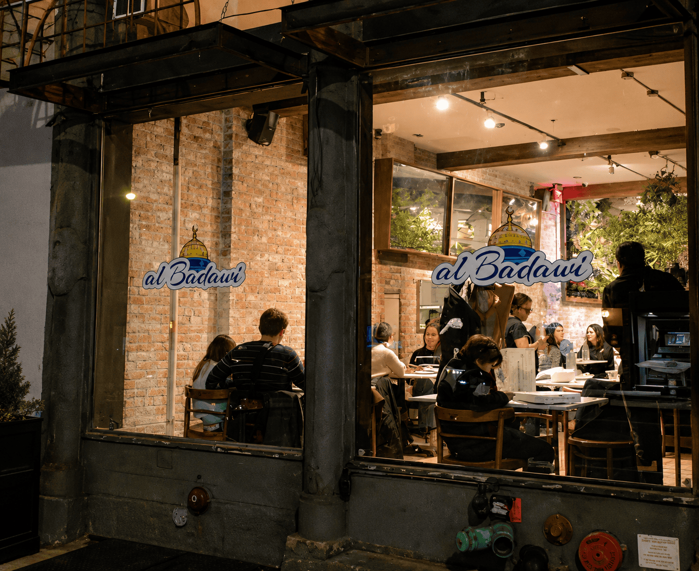 The storefront exterior of Al Badawi restaurant in Brooklyn at night, featuring large glass windows with the restaurant logo.