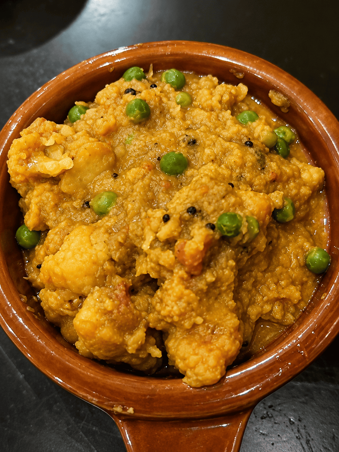 A close-up view of Aloo Cauli, a thick, golden-yellow potato and cauliflower curry mixed with bright green peas and black mustard seeds in a brown ceramic bowl.