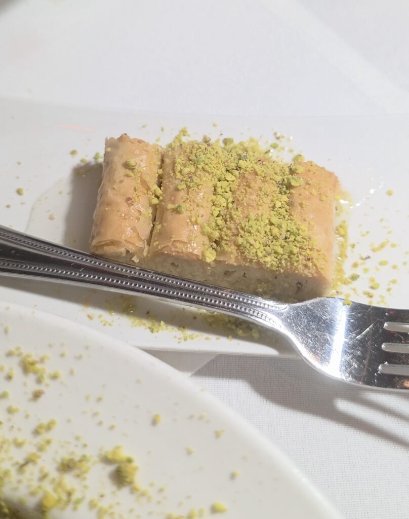 A piece of golden, layered filo pastry baklava on a small white plate, drenched in sweet syrup and coated with crushed green pistachios next to a metal fork.