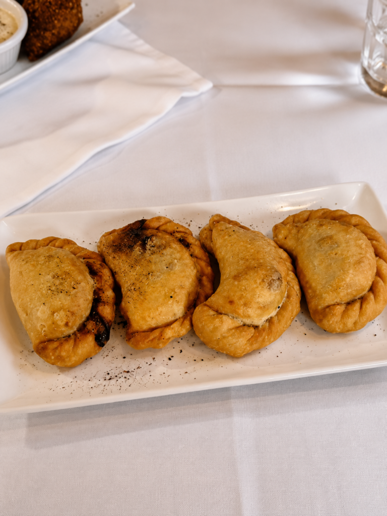 Four golden, crescent-shaped fried beef sambousek pastries lined up on a narrow white rectangular serving dish over a white tablecloth.