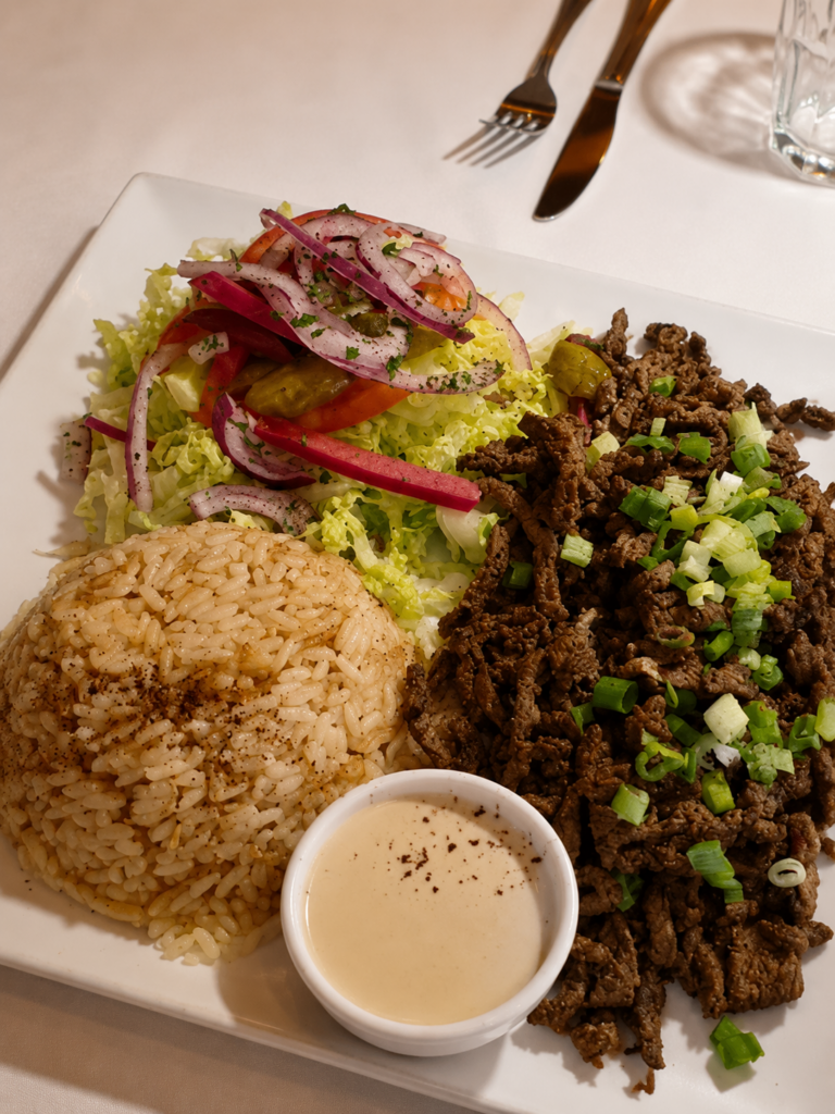 A close-up plate featuring a mountain of tender, thinly sliced beef shawarma topped with green onions, a scoop of spiced rice, a fresh side salad with red onions, and a small bowl of tahini sauce.