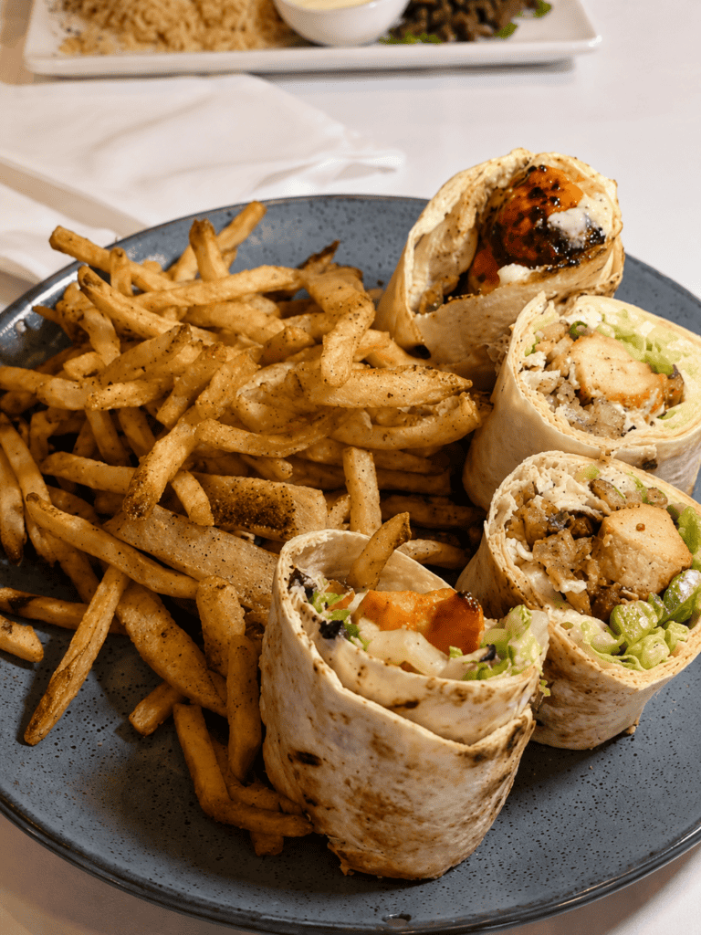 A blue round plate stacked high with golden French fries alongside four cross-sections of a chicken shish tawook wrap showing grilled chicken chunks, lettuce, and pickles.
