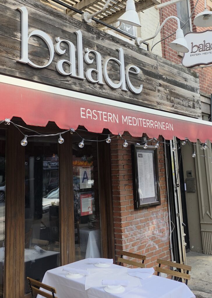 The exterior storefront of Balade restaurant in the East Village featuring its rustic wooden sign, red awning labeled Eastern Mediterranean, and an outdoor dining table set with white linen.