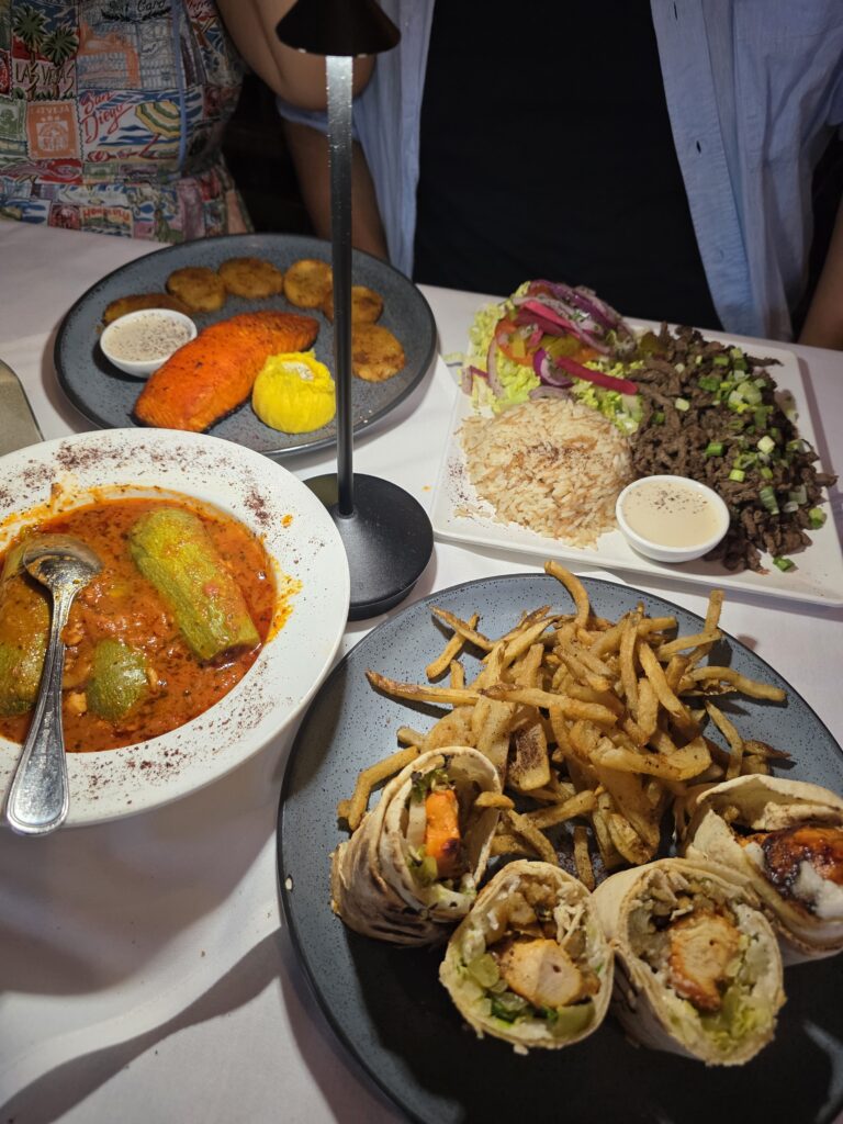 A full dinner table spread at Balade featuring plates of grilled salmon with potatoes, beef shawarma platter, stuffed kousa in tomato sauce, and a chicken wrap with fries under a table lamp.