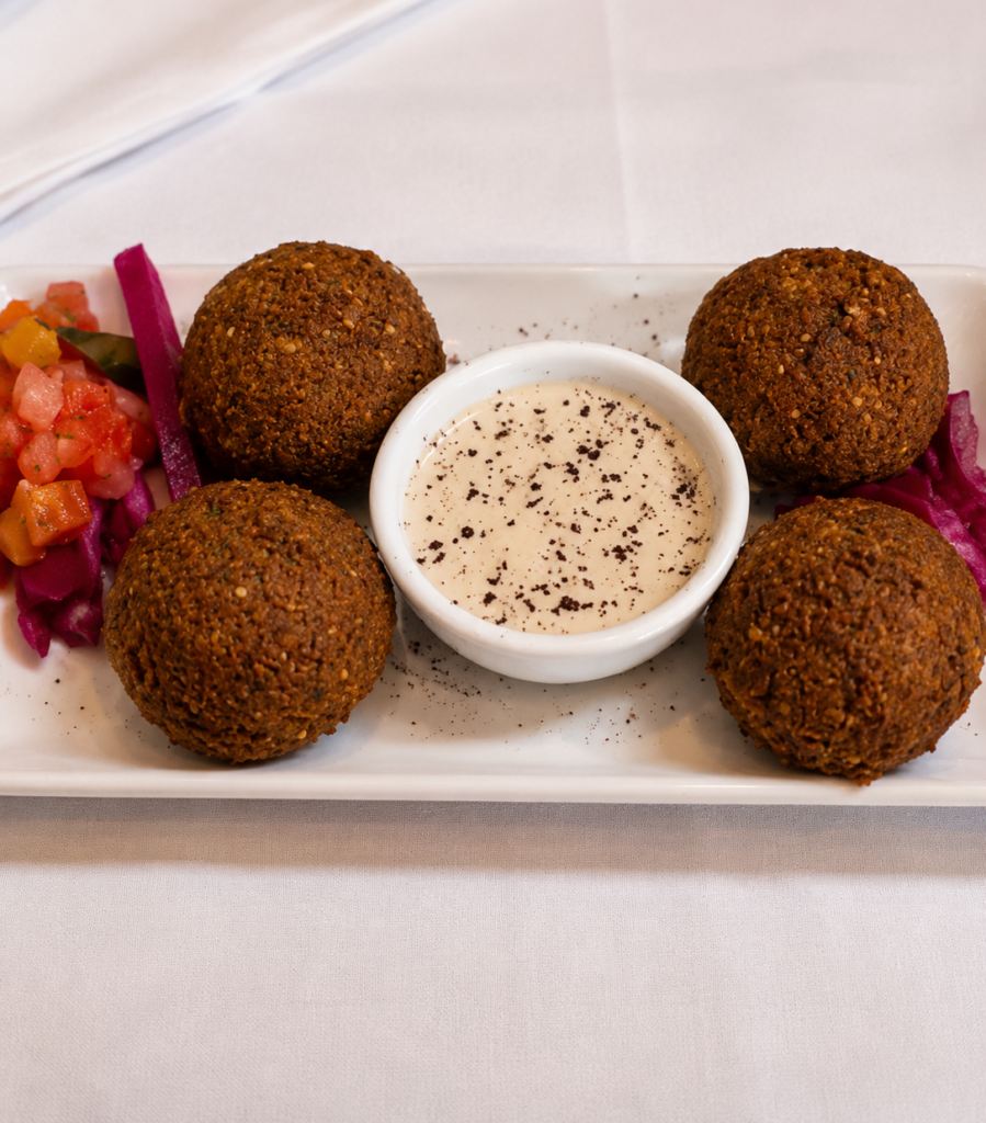 A top-down view of four golden-brown, crispy fried falafel balls served on a white rectangular plate with a central dish of creamy tahini sauce, pickled turnips, and diced tomatoes.