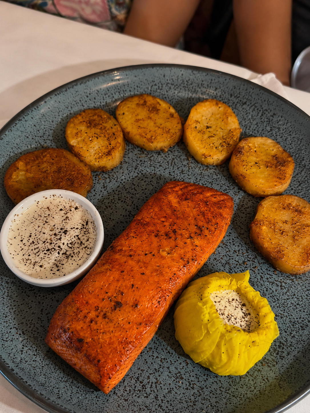 A speckled blue plate holding a large, perfectly seared rectangular salmon fillet, six seasoned roasted potato rounds arranged in a semi-circle, a small cup of white garlic sauce, and a half lemon wrapped in yellow cheesecloth.