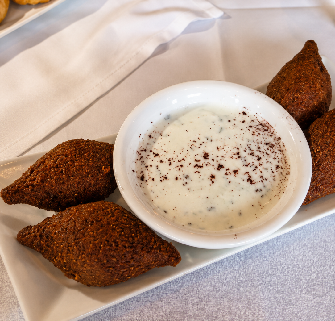 A white rectangular plate holding four golden-brown fried kibbeh croquettes arranged around a central white bowl filled with creamy laban yogurt sauce dusted with sumac spices.