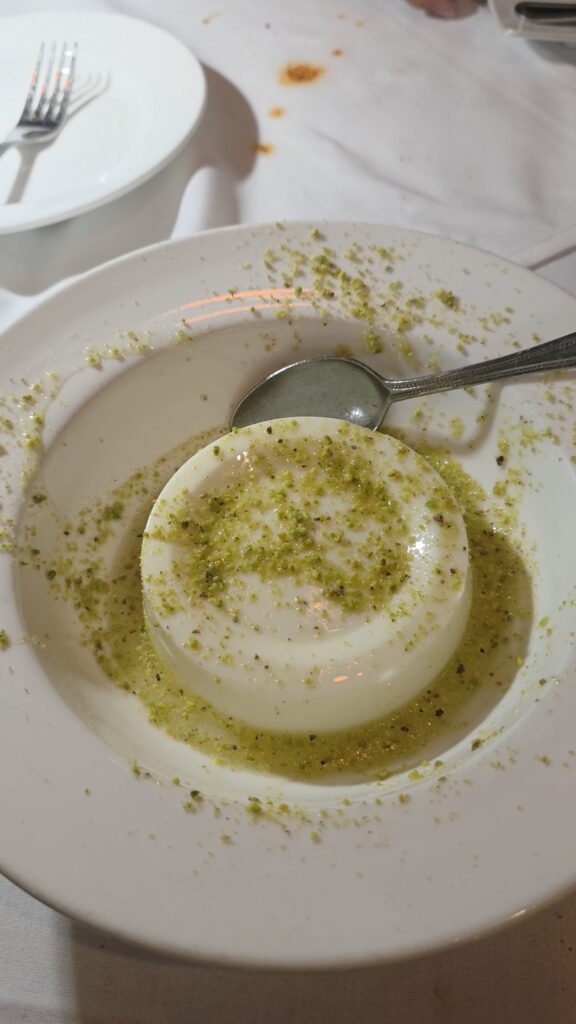 A round, smooth white Mouhallabie milk pudding sitting in a shallow white dessert bowl, generously topped and surrounded by bright green crushed pistachios and syrup.