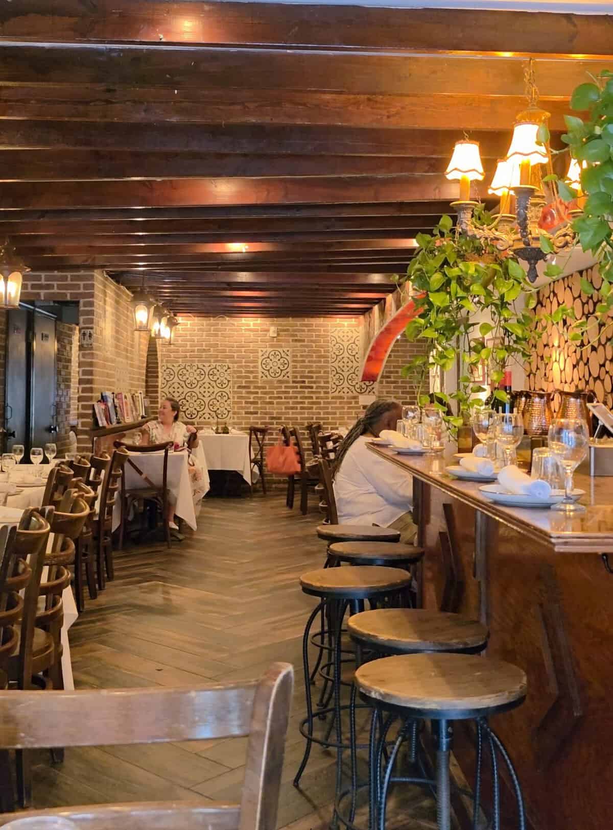 The warm interior dining room of Balade featuring dark exposed wooden ceiling beams, brick walls with patterned accent tiles, a wooden bar with stools, and tables set with white tablecloths.