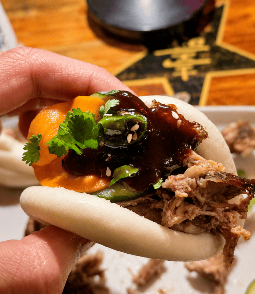 A hand holding an assembled steamed bao bun packed with shredded duck meat, hoisin sauce, sliced pickles, and fresh cilantro.