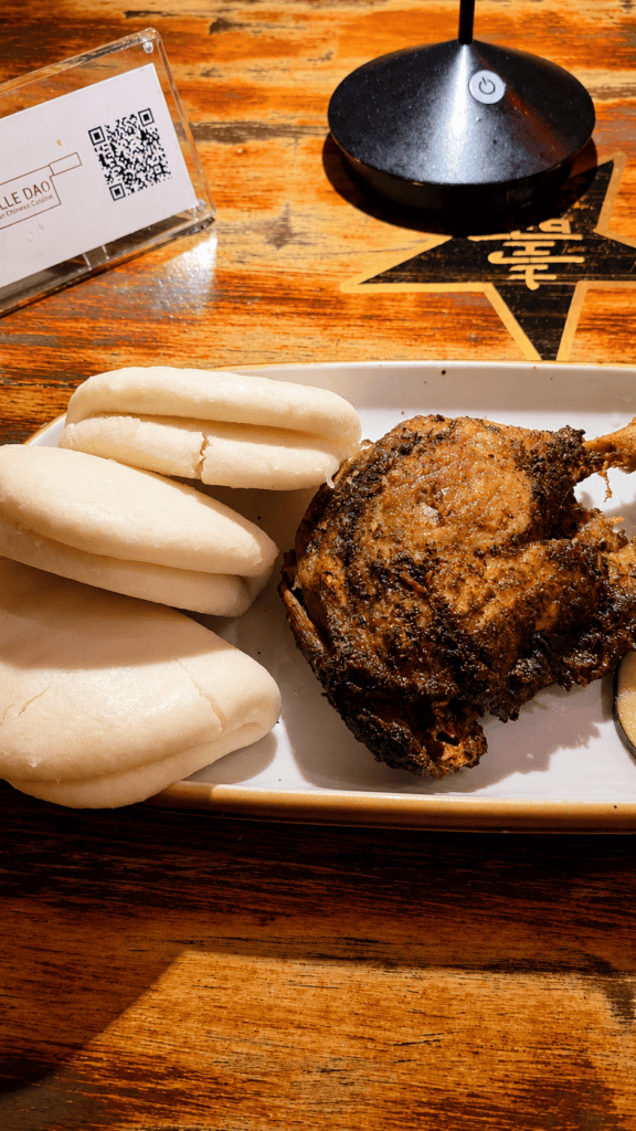 A plate featuring crispy roasted duck leg alongside a stack of soft white steamed bao buns.