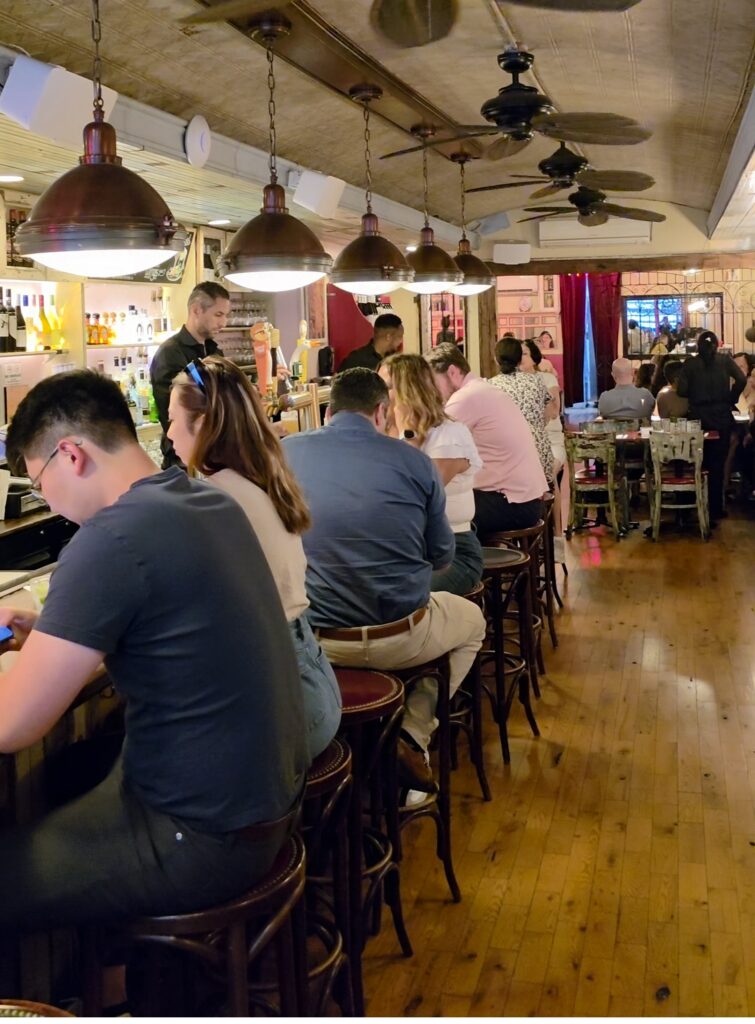 A lively, warmly lit restaurant bar counter with diners seated on wooden barstools beneath copper pendant lamps.