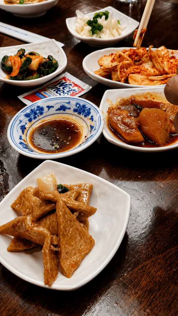 An array of complimentary Korean banchan side dishes on a dark wooden table including cabbage kimchi, fish cakes, seasoned cucumber, pickled green beans, and savory potatoes.